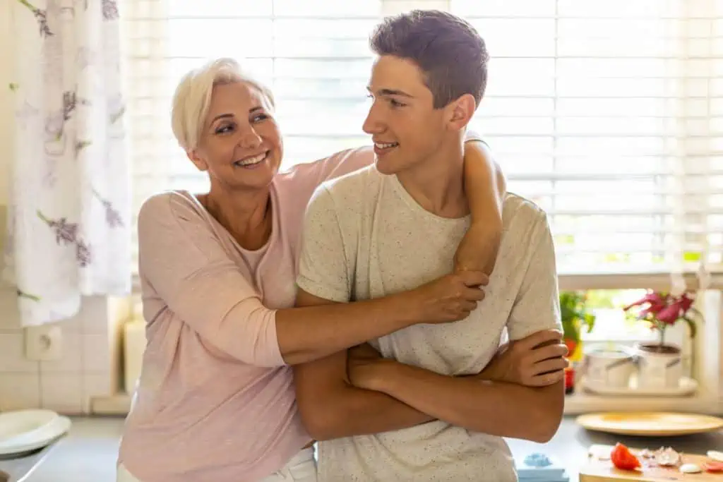 Smiling mother with her arm around her teenage son in a bright kitchen, sharing a moment of connection.